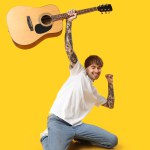 Young tattooed man with guitar dancing against yellow background