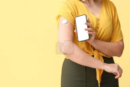 Frau mit Glukosesensor zur Messung des Blutzuckerspiegels und Smartphone auf gelbem Hintergrund. Diabetes-Konzept