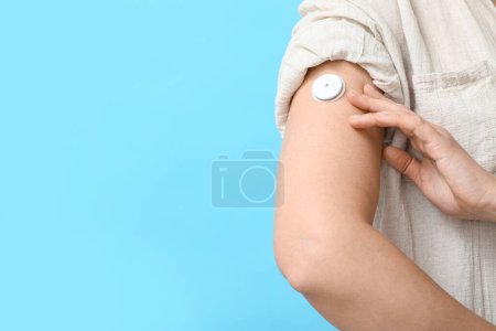 Téléchargez les photos : Femme avec capteur de glucose pour mesurer le taux de sucre dans le sang sur fond bleu, gros plan. Concept de diabète - en image libre de droit