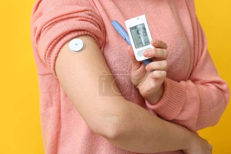 Woman with glucometer, lancet pen and sensor for measuring blood sugar level on yellow background, closeup. Diabetes concept