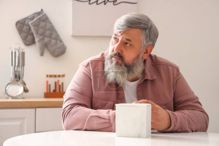 Portrait of sad senior man with tissues sitting at table in kitchen