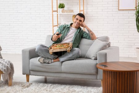Photo for Young man holding cardboard box with tasty pizza and TV remote control on sofa at home - Royalty Free Image