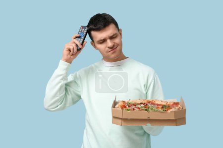 Photo for Young man holding cardboard box with tasty pizza and TV remote control on blue background - Royalty Free Image
