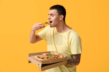 Photo for Young man eating piece of tasty pizza and holding cardboard box on yellow background - Royalty Free Image