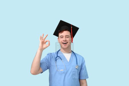 Photo for Male student in mortar board and uniform of doctor showing ok gesture on blue background - Royalty Free Image