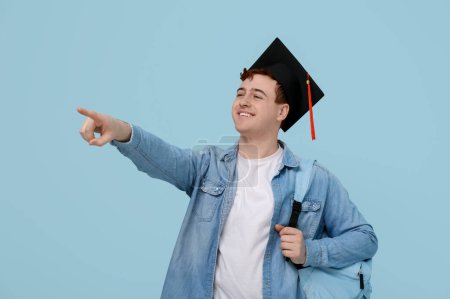 Photo for Male student in mortar board with backpack pointing at something on blue background - Royalty Free Image