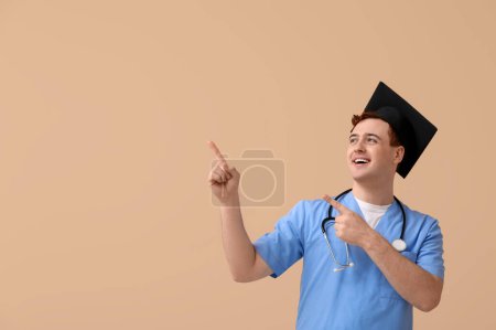 Photo for Male student in mortar board and uniform of doctor pointing at something on beige background - Royalty Free Image