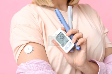 Femme avec glucomètre, capteur pour mesurer le taux de sucre dans le sang et stylos à lancettes sur fond rose, gros plan. Concept de diabète