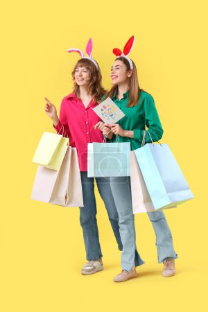 Mujeres jóvenes felices en orejas de conejo de Pascua diademas con bolsas de compras de papel y postal festiva sobre fondo amarillo