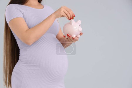 Young pregnant woman putting coin into piggy bank on light background, closeup. Maternity Benefit concept