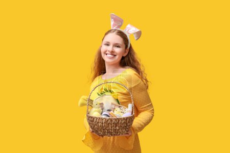 Schöne junge glückliche Frau in Hasenohren mit Weidenkorb voller dekorativer Kosmetik auf gelbem Hintergrund