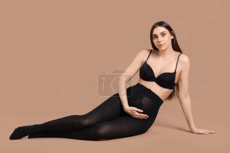 Junge schwangere Frau in schwarzen Strumpfhosen sitzt auf beigem Hintergrund