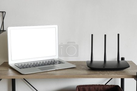 Modern wi-fi router with blank laptop on shelf near light wall