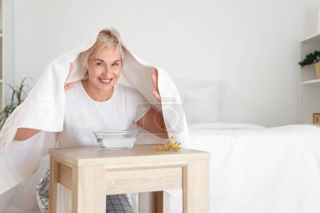 Mature woman doing steam inhalation at table in bedroom