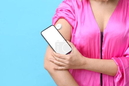 Woman with glucose sensor for measuring blood sugar level and phone on blue background, closeup. Diabetes concept