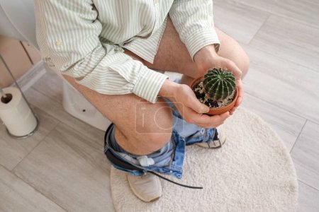 Young man with cactus sitting on toilet bowl in restroom, closeup. Hemorrhoids concept
