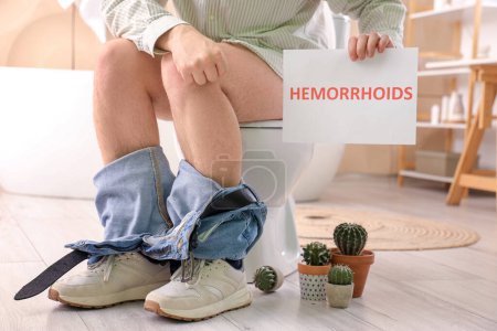 Young man with cacti sitting on toilet bowl in restroom. Hemorrhoids concept