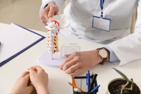 Médico masculino explicando anatomía espinal con modelo de columna vertebral al paciente en clínica, primer plano