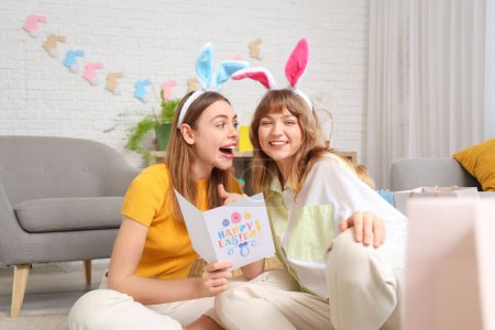Mujeres jóvenes felices en orejas de conejo diademas con bolsas de compras de papel y postal festiva para Pascua en casa