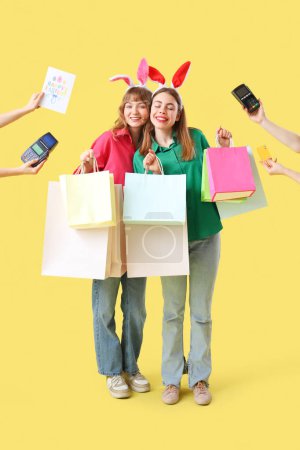 Glücklich lächelnde junge Frauen mit Hasenohren-Stirnbändern mit Papiereinkaufstaschen für Ostern und Händen mit Zahlungsterminals auf gelbem Hintergrund