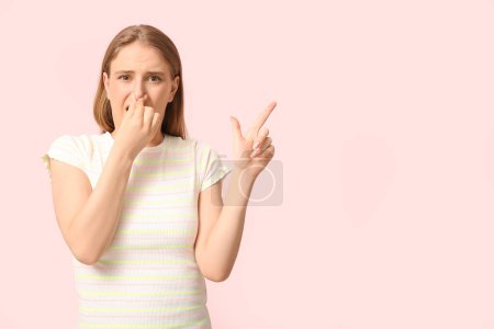 Young woman feeling disgusting smell and pointing at something on pink background