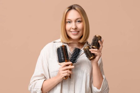 Beautiful young woman with bob hairstyle holding hair brushes on brown background