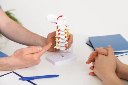 Médico masculino explicando anatomía espinal con modelo de columna vertebral al paciente en clínica, primer plano