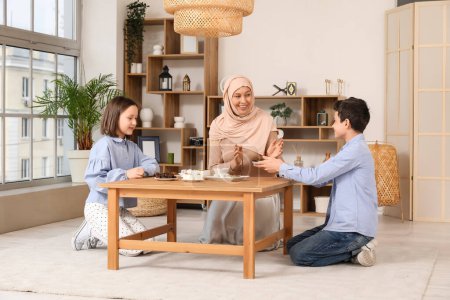 Glückliche muslimische Mutter und ihre Kinder sitzen mit traditionellen Süßigkeiten im Wohnzimmer am Tisch. Eid al-Fitr-Feier