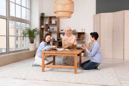 Happy Muslim mother and her children sitting at table with traditional sweets in living room. Eid al-Fitr celebration