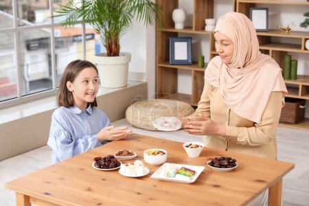 Happy Muslim mother and her daughter sitting at table with traditional sweets in living room. Eid al-Fitr celebration