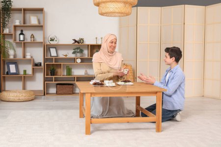 Glückliche muslimische Mutter und ihr Sohn sitzen mit traditionellen Süßigkeiten im Wohnzimmer am Tisch. Eid al-Fitr-Feier