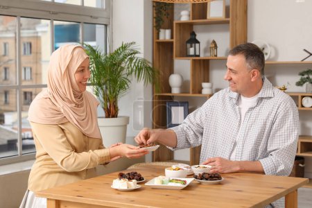 Glückliche Muslimin mit ihrem Mann am Tisch mit traditionellen Süßigkeiten im Wohnzimmer. Eid al-Fitr-Feier