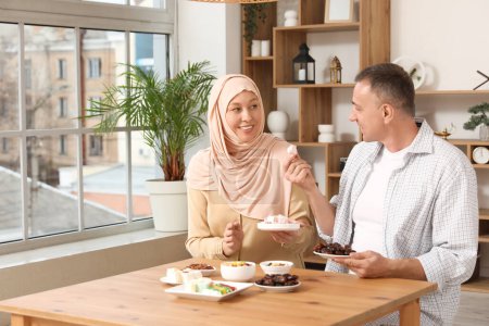 Glückliche Muslimin mit ihrem Mann am Tisch mit traditionellen Süßigkeiten im Wohnzimmer. Eid al-Fitr-Feier