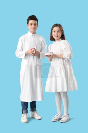 Cute Muslim kids with traditional sweets on blue background. Eid al-Fitr celebration