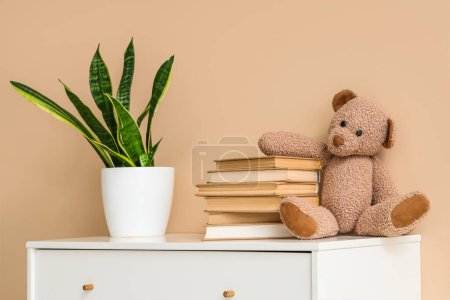 Foto de Cómoda con libros, oso de juguete y planta de interior cerca de la pared beige - Imagen libre de derechos
