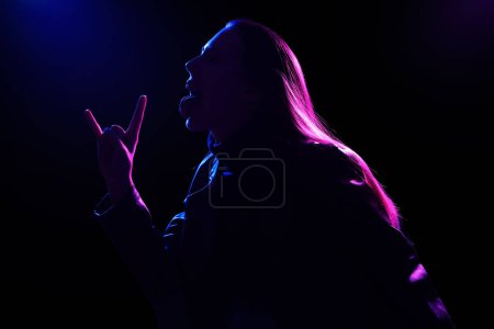 Téléchargez les photos : Silhouette de jeune femme montrant "cornes du diable" sur fond sombre - en image libre de droit