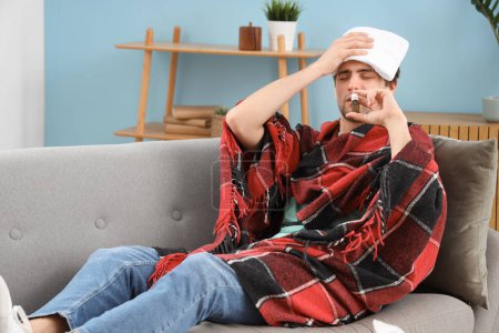 Sick young man with towel using nasal drops on sofa at home