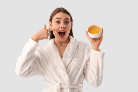 Schöne junge schockierte Frau im Bademantel zeigt auf Container mit Zuckerpaste auf grauem Hintergrund