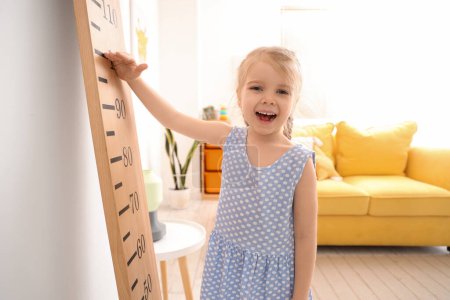Cute little girl measuring height near wooden stadiometer at home