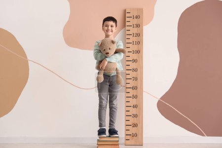 Cute little boy on books measuring height near wooden stadiometer at home