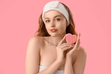 Beautiful young woman in bath headband with heart-shaped sponge on pink background, closeup