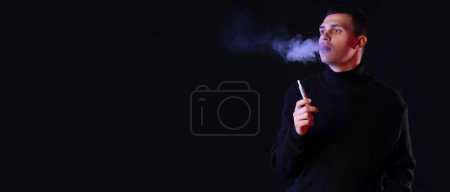 Téléchargez les photos : Jeune homme fumant cigarette électronique sur fond noir avec espace pour le texte - en image libre de droit