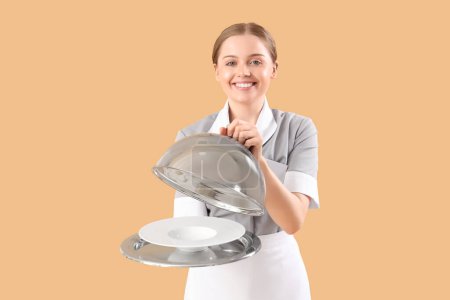 Téléchargez les photos : Jeune serveuse avec assiette vide, plateau et cloche sur fond beige - en image libre de droit