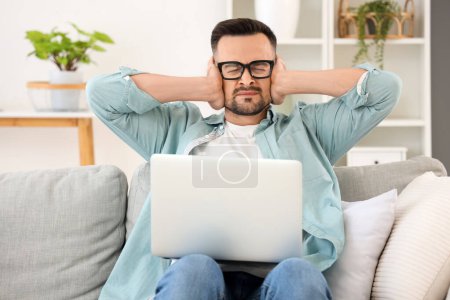Working young man with laptop suffering from loud neighbours at home