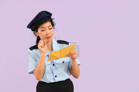 Young Asian postwoman with poor eyesight reading address on envelope against lilac background