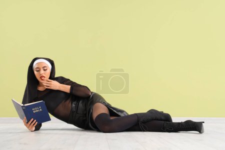 Sexy nun with Holy Bible licking finger near green wall