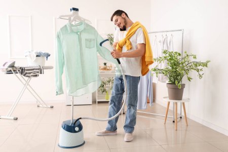 Happy young man steaming green shirt and talking by mobile phone in laundry room