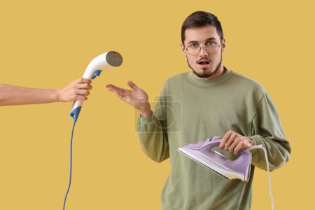 Shocked young man with iron and hand holding modern steamer on yellow background