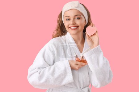 Beautiful young woman in bath headband with heart-shaped sponge and serum on pink background