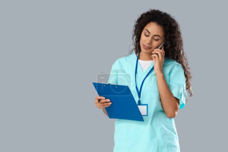 Female African-American medical intern with clipboard talking by mobile phone on light background